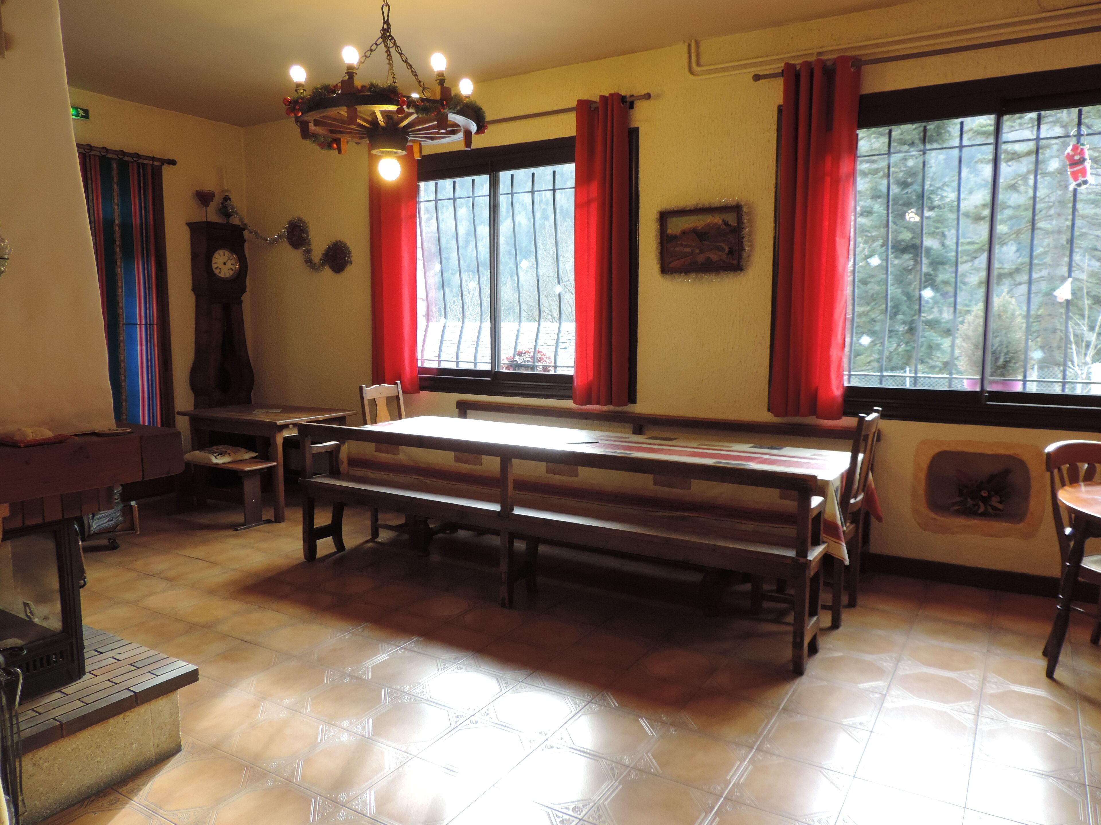 Un comedor con una larga mesa de madera, bancos, un reloj y un ventanal con cortinas rojas.