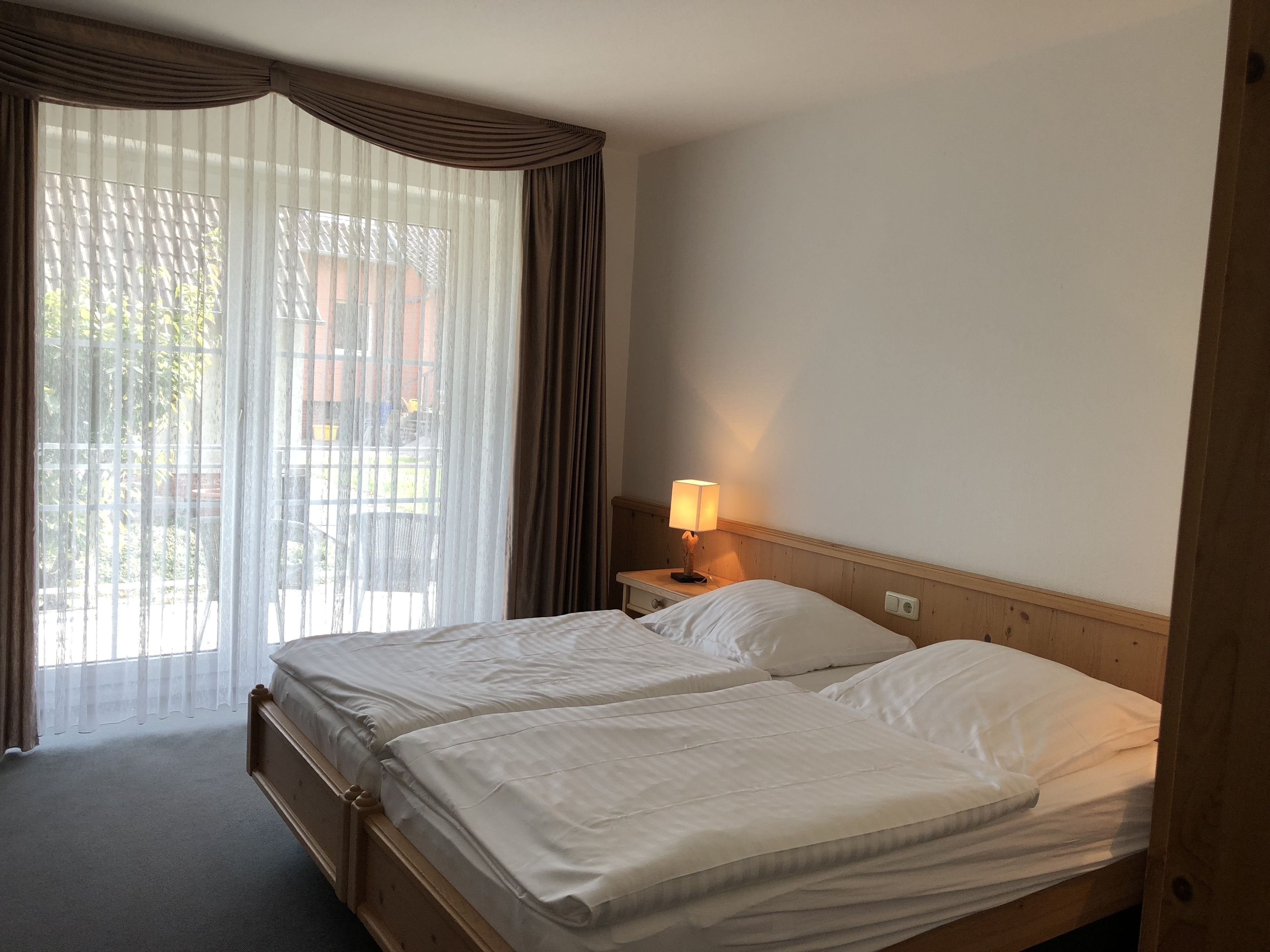 Habitación de hotel con dos camas, una mesita de noche con lámpara y vistas al exterior a través de cortinas traslúcidas.