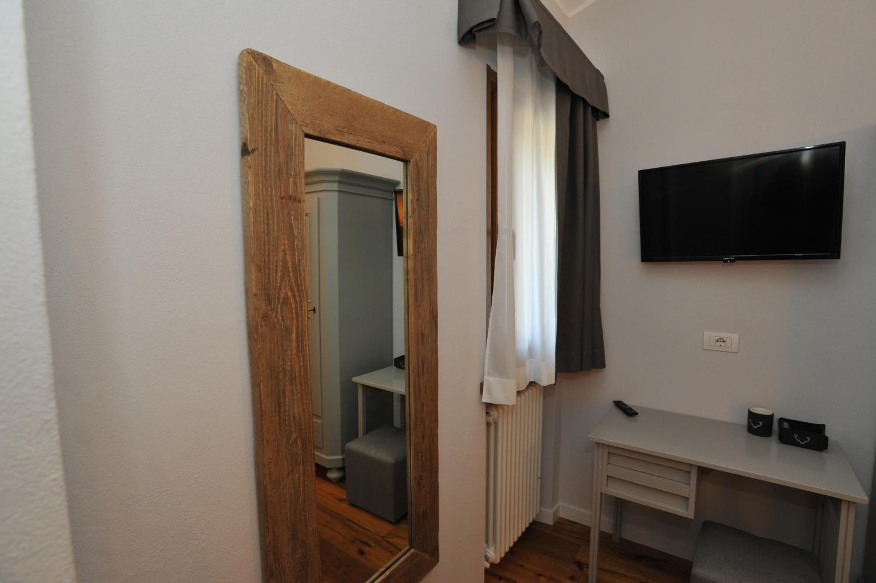 A room with a wooden door, a mirror, a radiator, a TV mounted on the wall, a desk, and a chair.