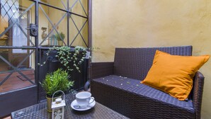 A wicker sofa with a single orange cushion, a small table with a cup and saucer, and a potted plant. - Florence Ariento Romantic Apartment (Florence)