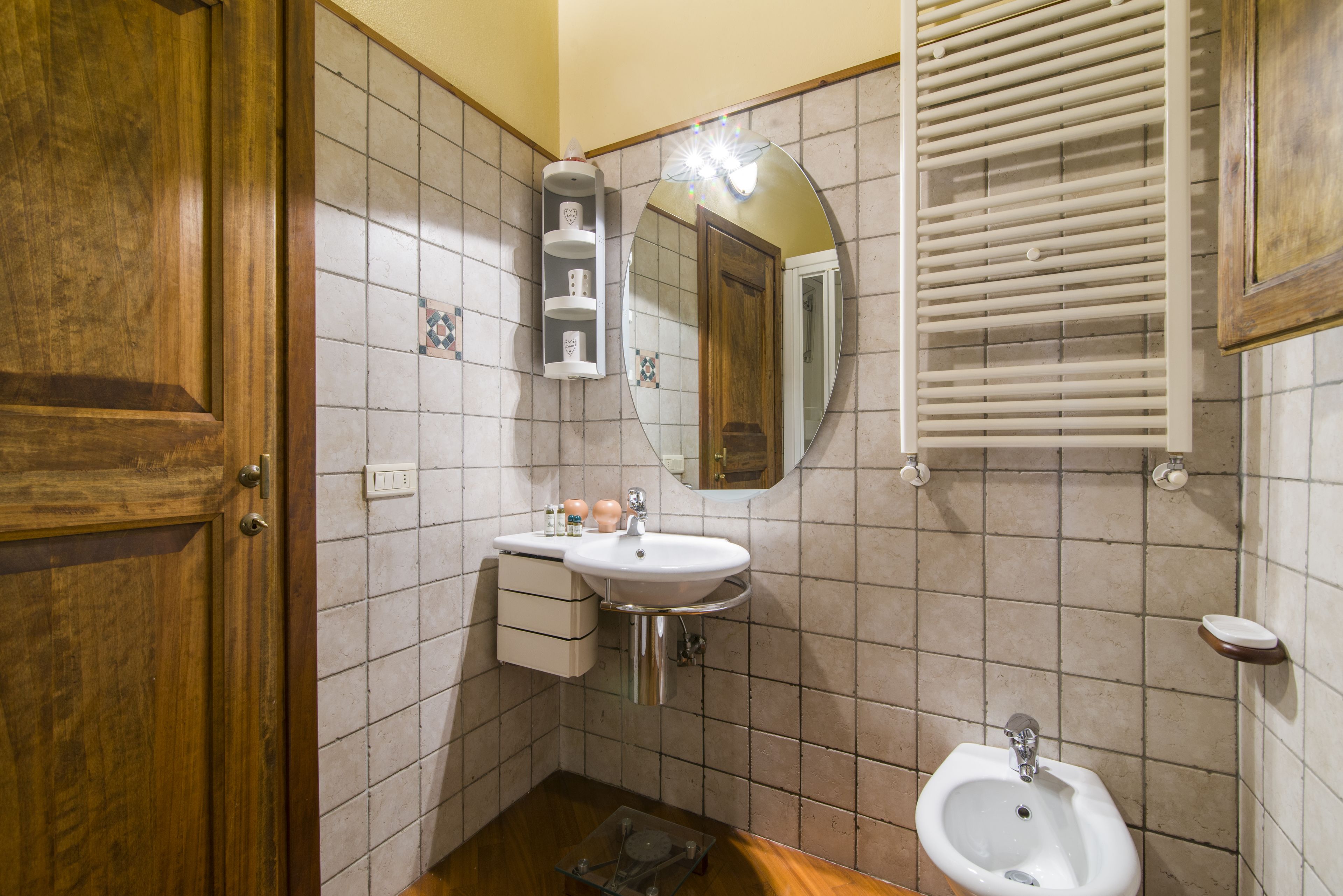 A bathroom with a wooden door, a white sink with a mirror above it, a bidet, and a tiled wall.