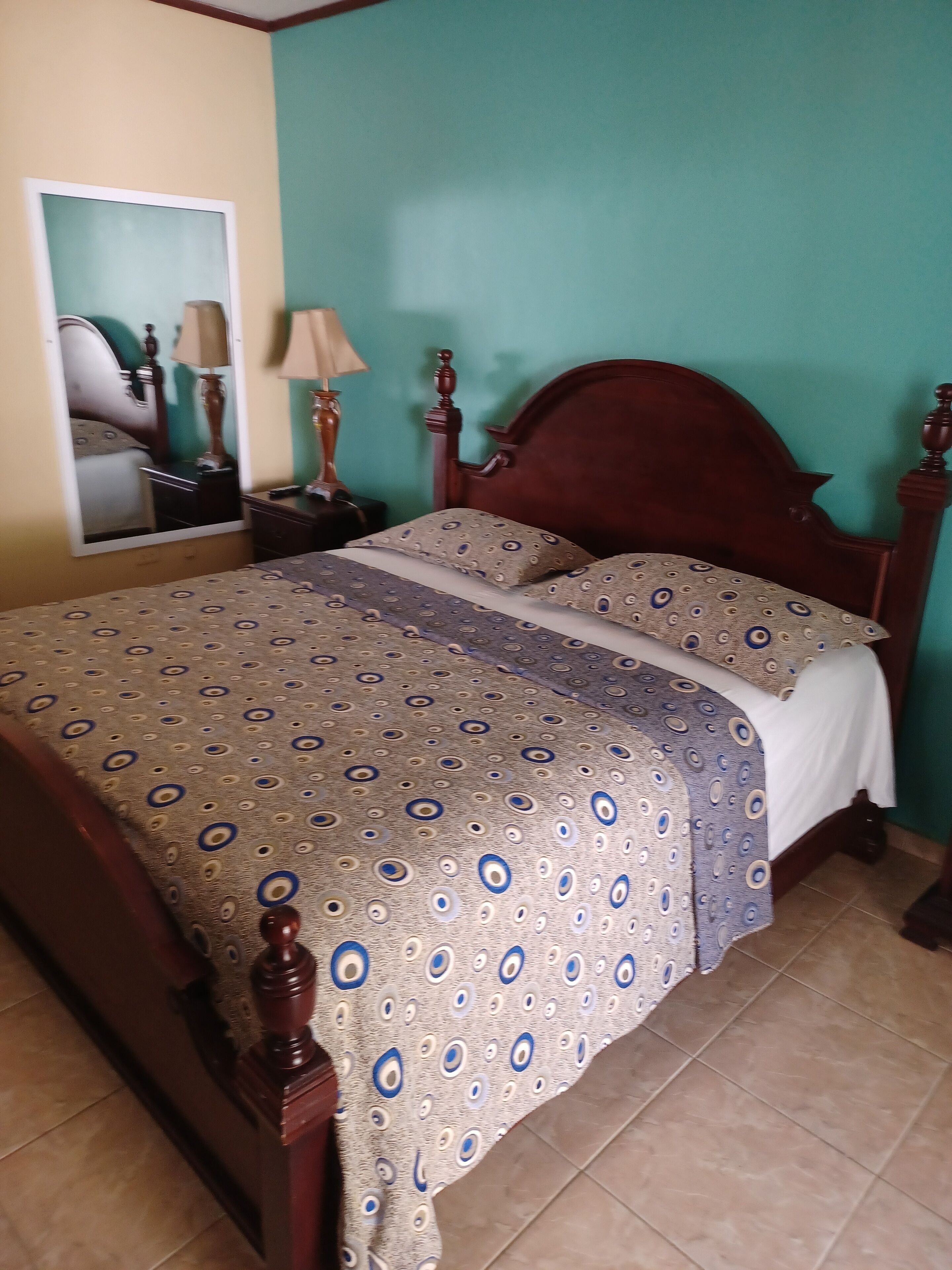 A bedroom with a wooden bed, patterned bedspread, a mirror, and a nightstand with a lamp.
