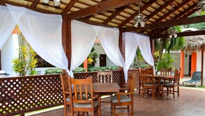 An outdoor patio with wooden tables and chairs, a thatched roof, and white drapes. - Oasis Adult Resort (Puerto Plata)