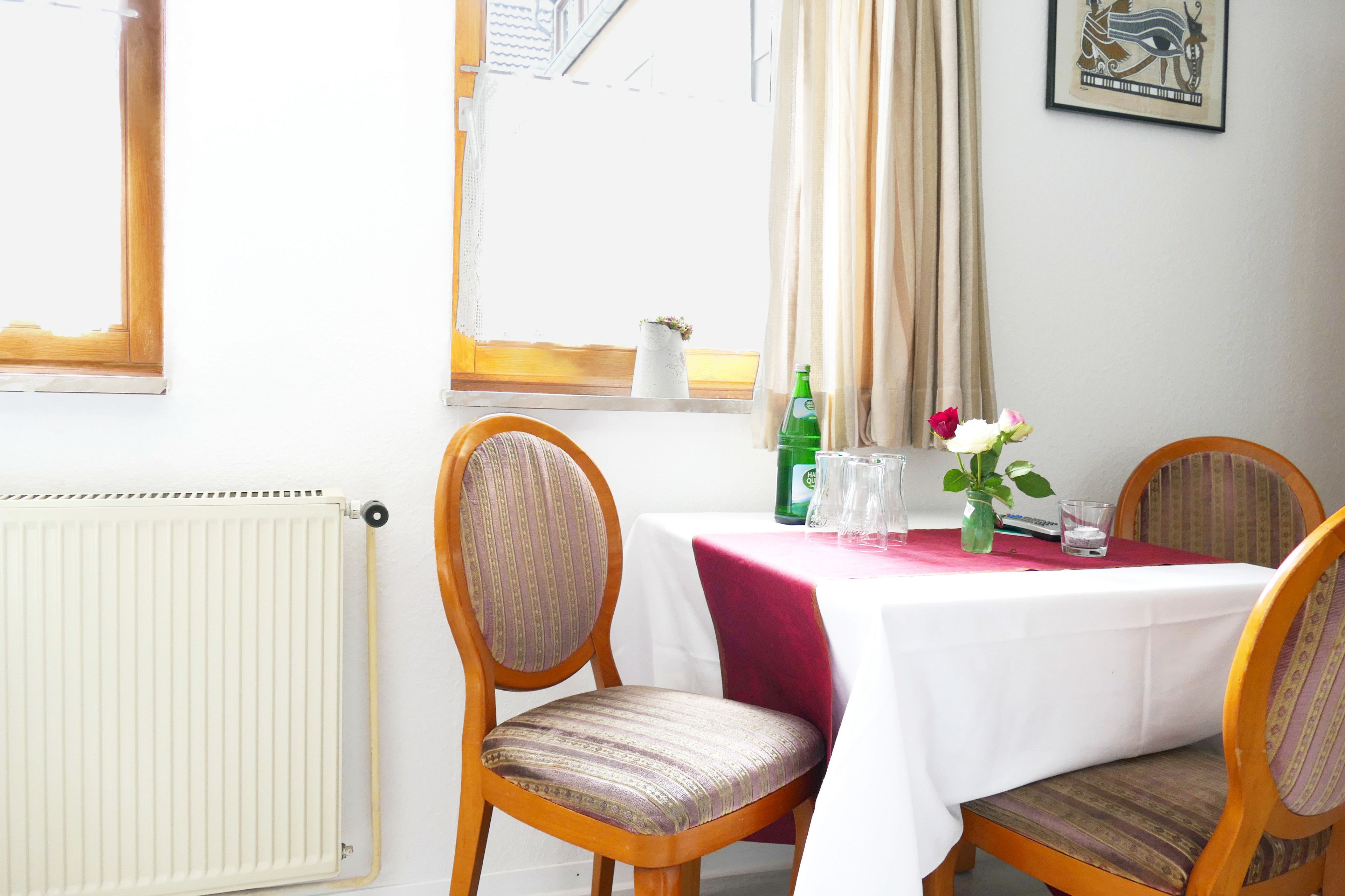A dining area with a table set for two, chairs, a radiator, a window with curtains, and a framed picture on the wall.