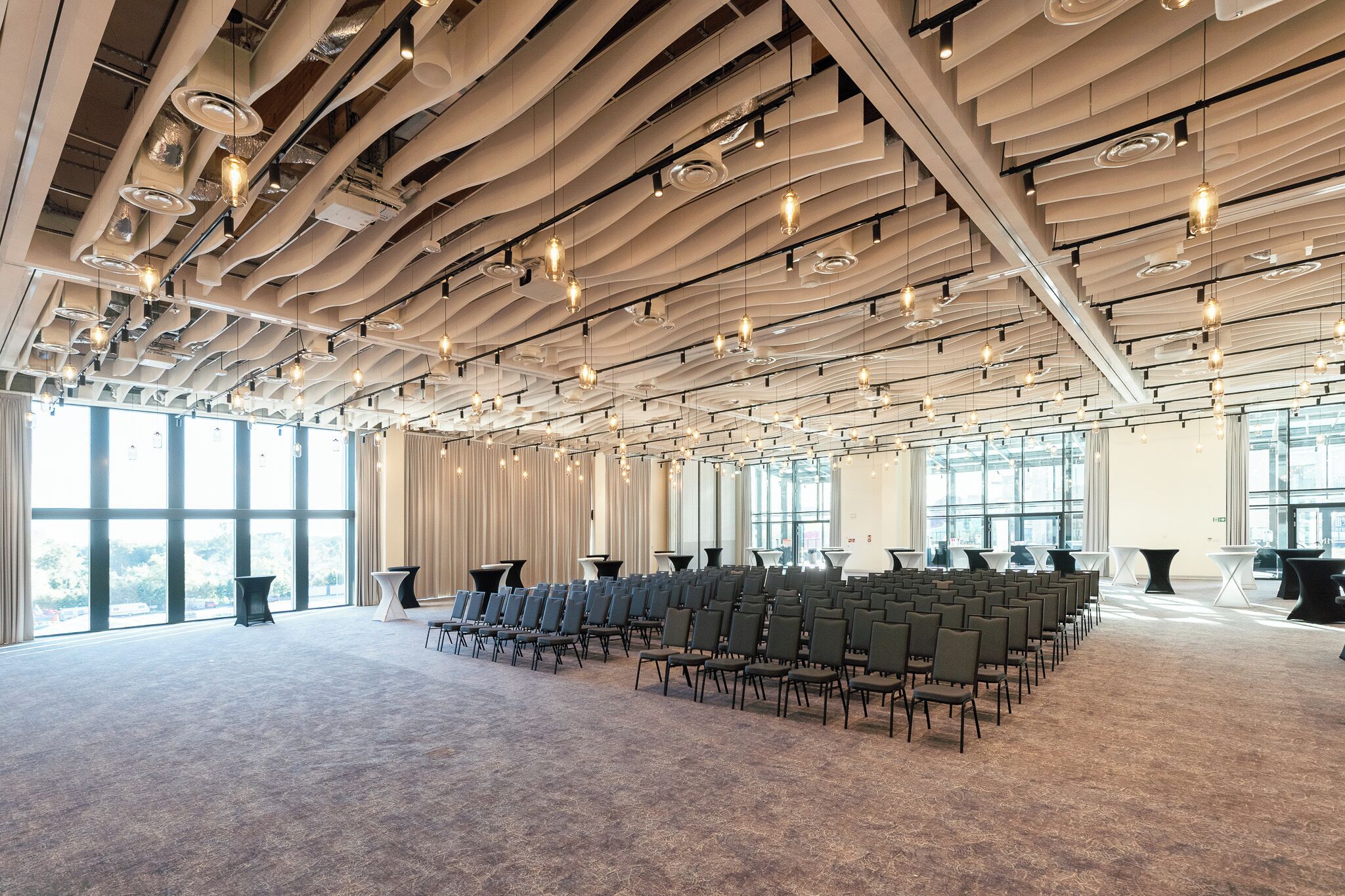 Una amplia sala de conferencias con filas de sillas, ventanas grandes y un diseño de techo moderno.