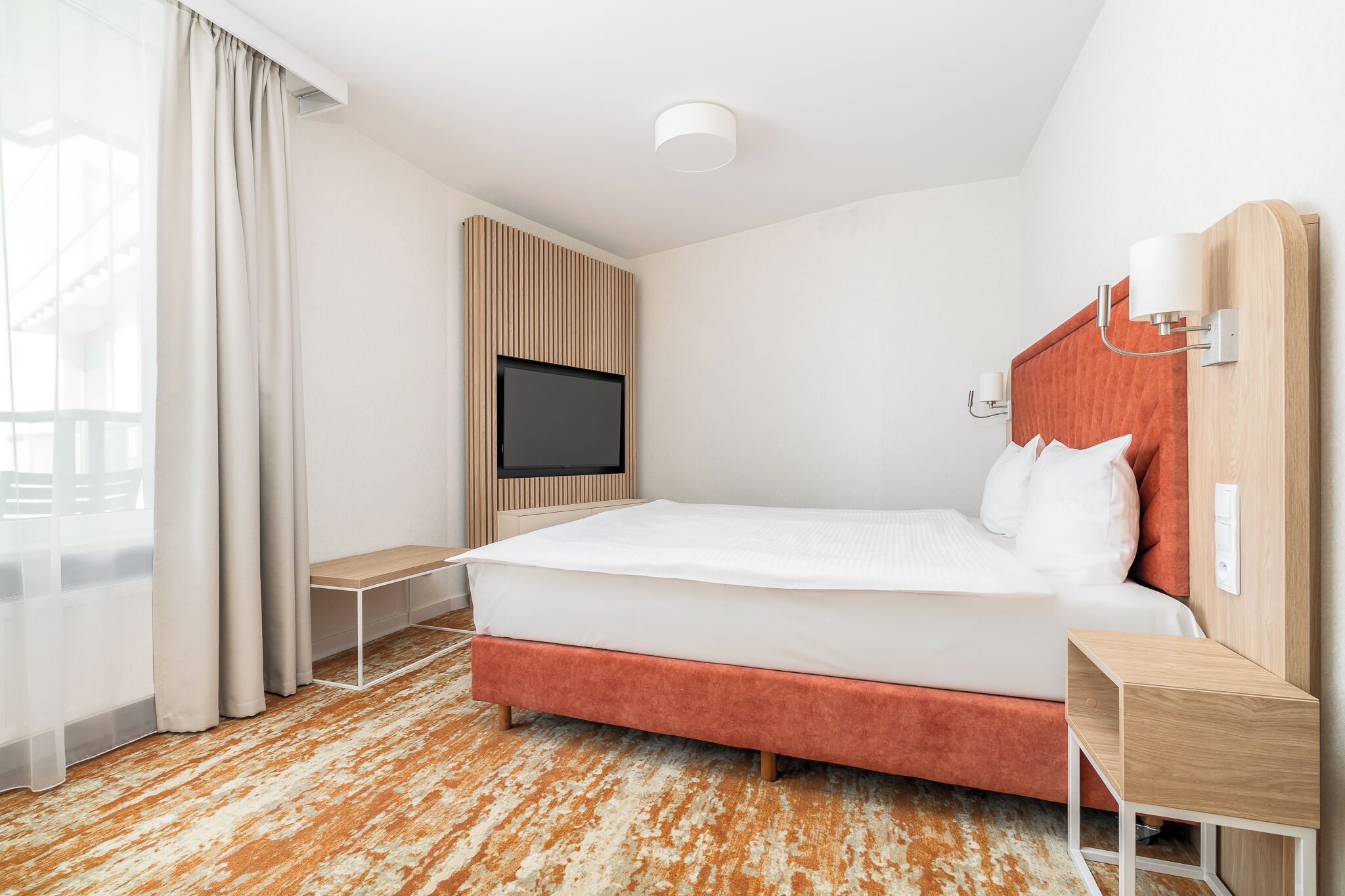 A modern hotel room with a large bed, a TV mounted on the wall, a wooden headboard, and a small bedside table.