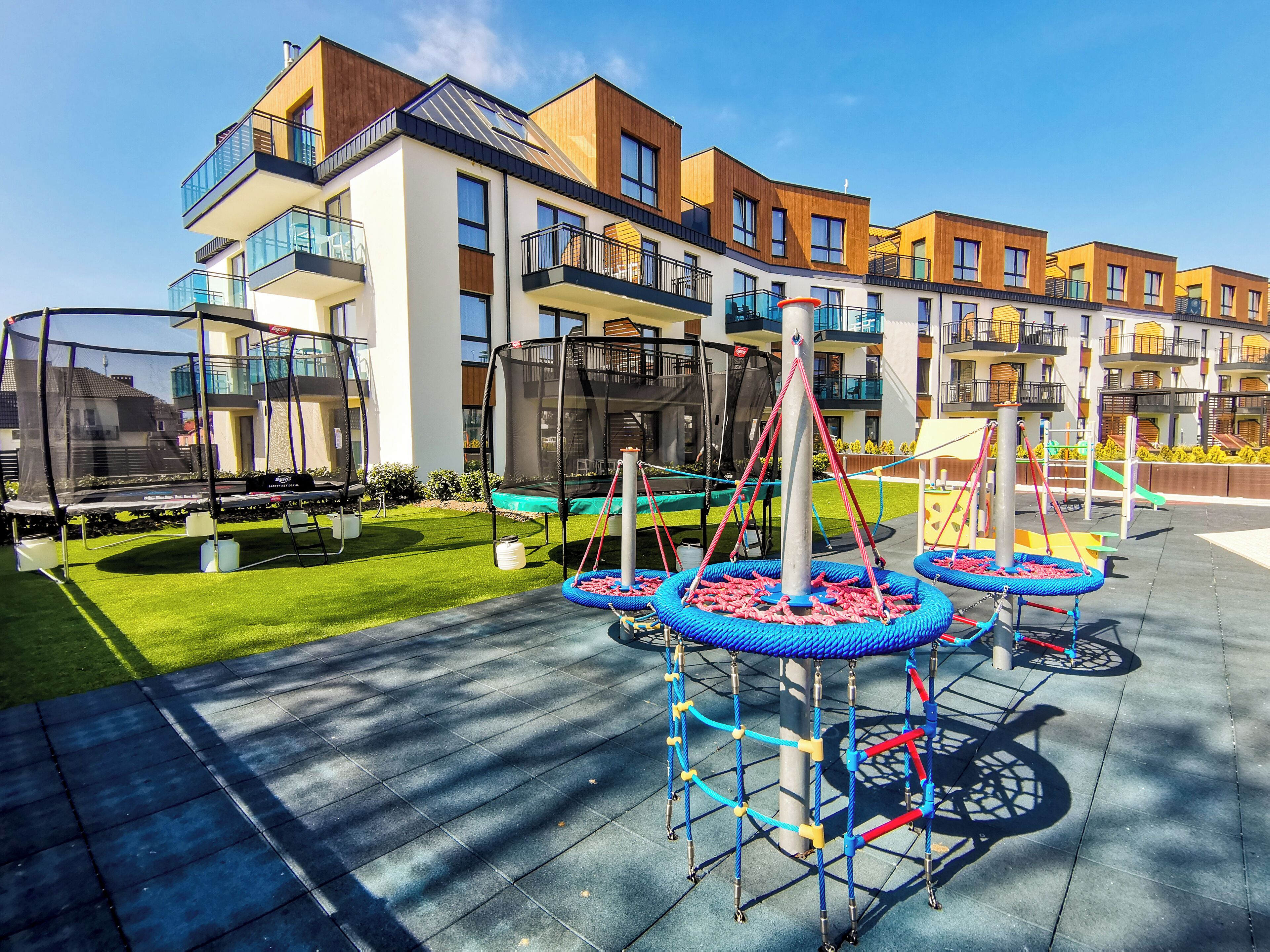 A playground with a trampoline, a swing set, and a climbing structure in front of a modern apartment building.