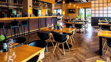 A restaurant with wooden tables and black chairs, a bar area with shelves of bottles and glasses, and a herringbone patterned floor.