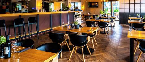 A restaurant with wooden tables and black chairs, a bar area with shelves of bottles and glasses, and a herringbone patterned floor.