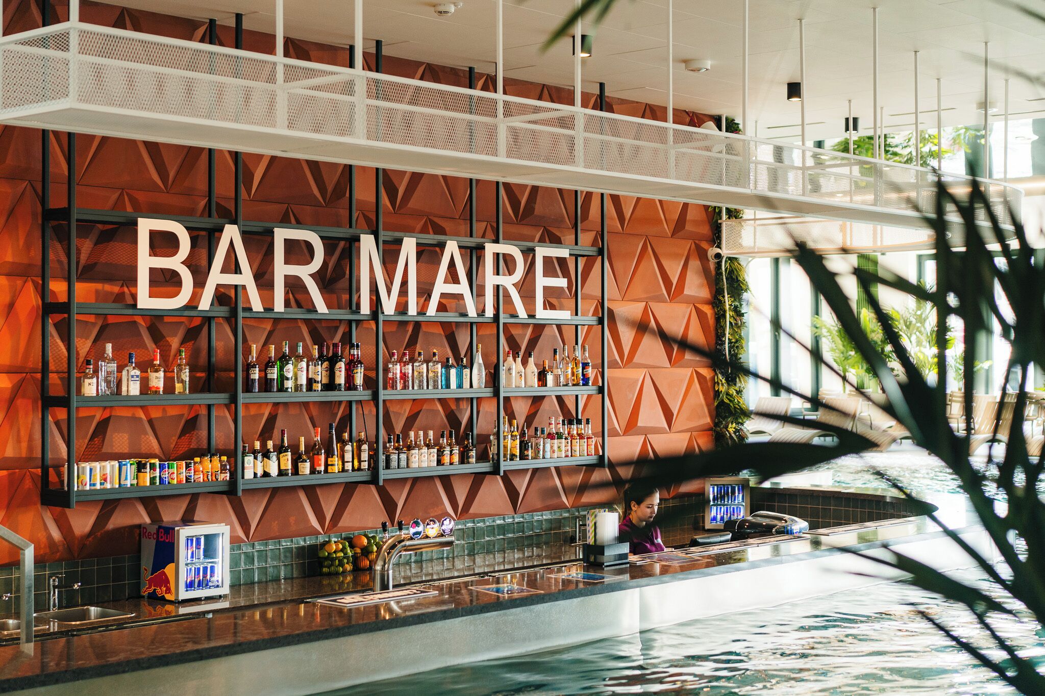 A bar area with shelves of liquor bottles, a Red Bull cooler, and a pool in the background.