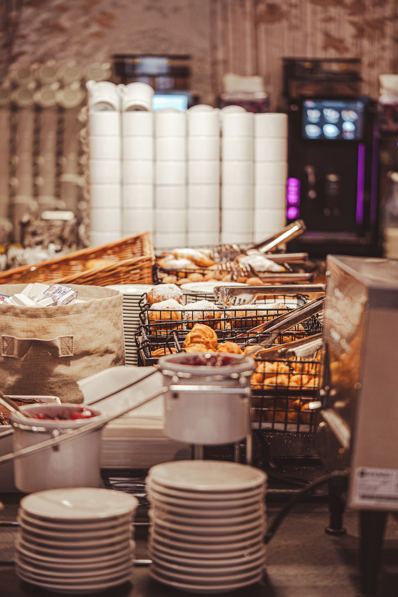 A buffet setup with various food items in baskets, stacks of plates, and a coffee machine in the background.