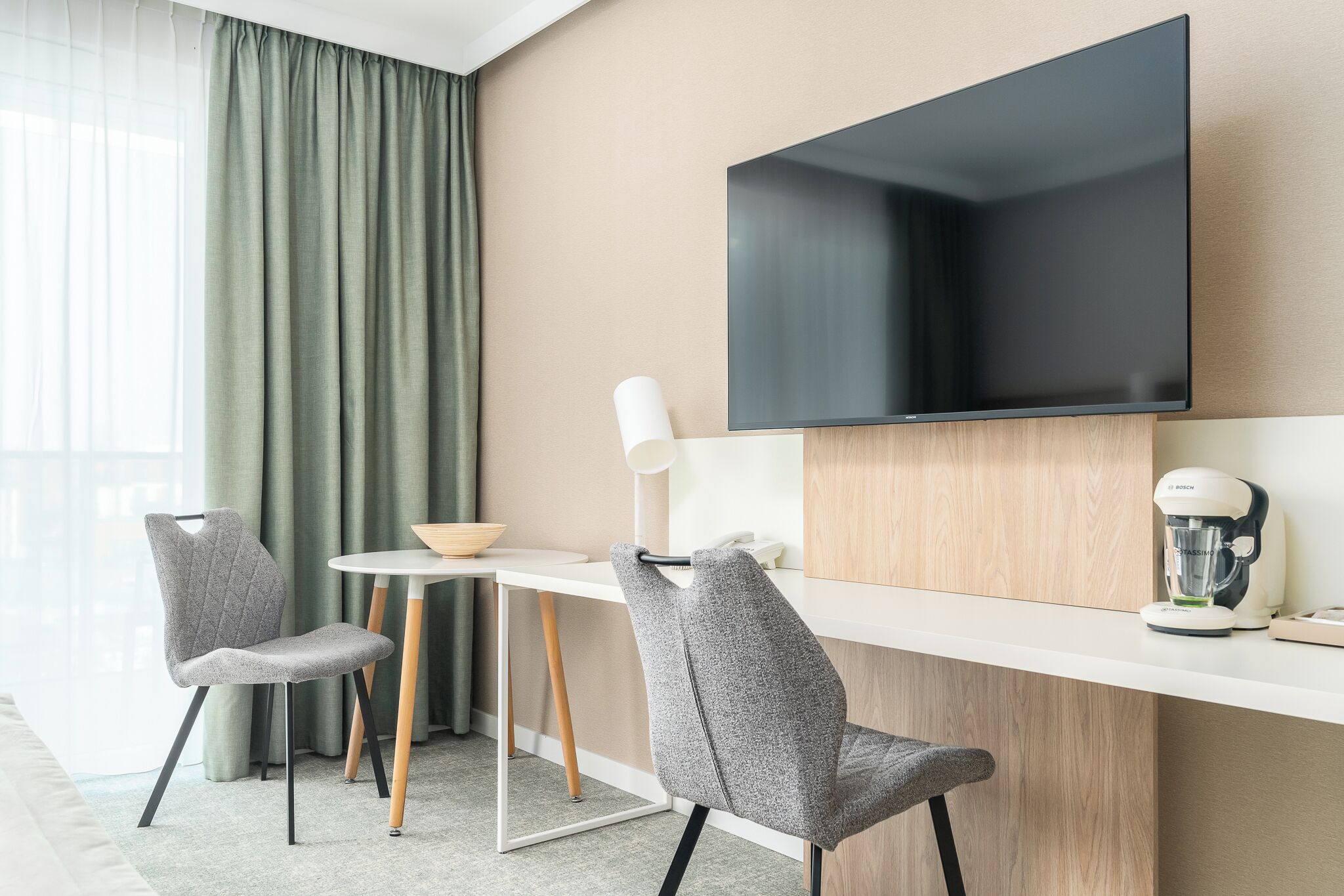 A modern hotel room with a flat-screen TV mounted on the wall, a small round table with a bowl, two grey chairs, a white lamp, and a coffee maker on a wooden counter.
