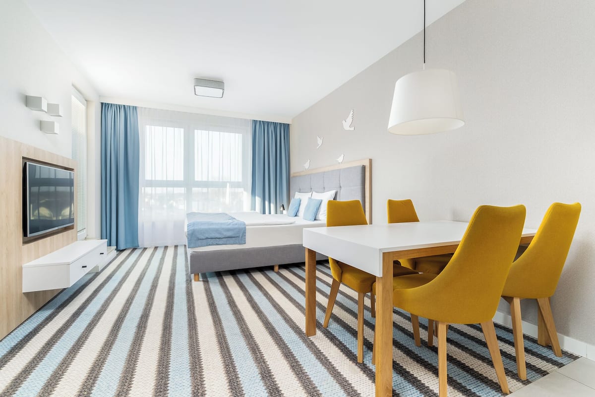 Une chambre d’hôtel moderne équipée d’un lit, d’une télévision, d’une table à manger avec des chaises et d’une grande fenêtre avec des rideaux.