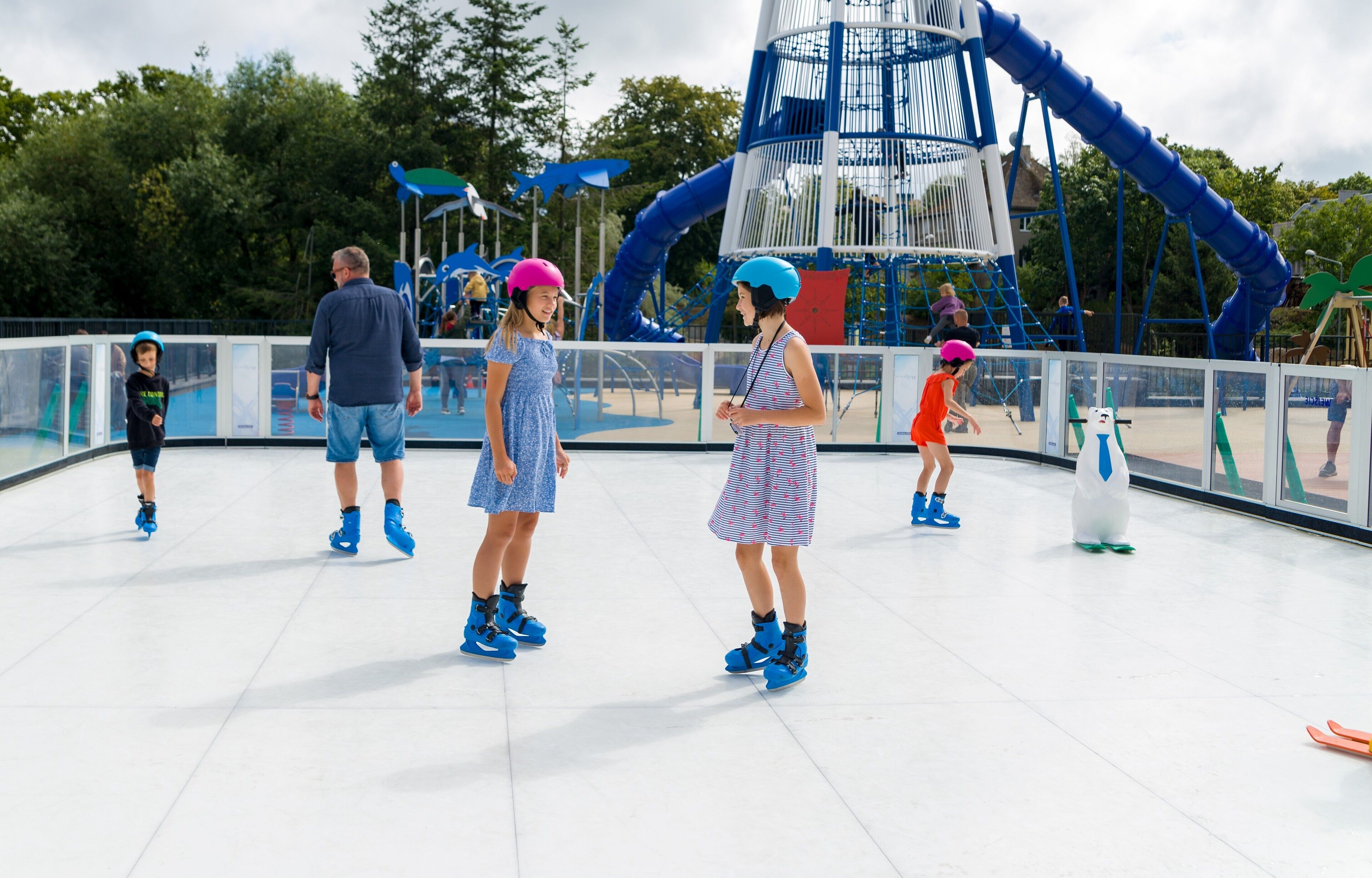 A group of children roller skating on a smooth, white surface with a large blue slide structure in the background.