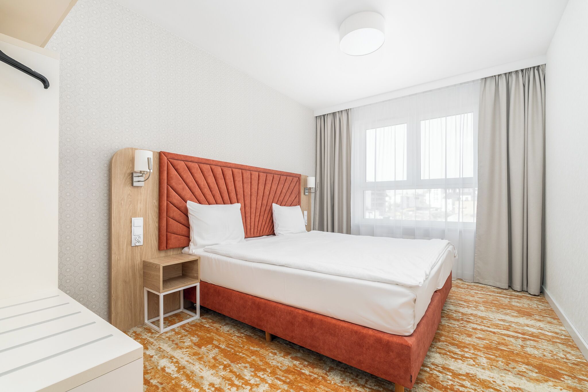 A modern hotel room with a large bed, a wooden nightstand, a white bedside table, and a view of the city through large windows.