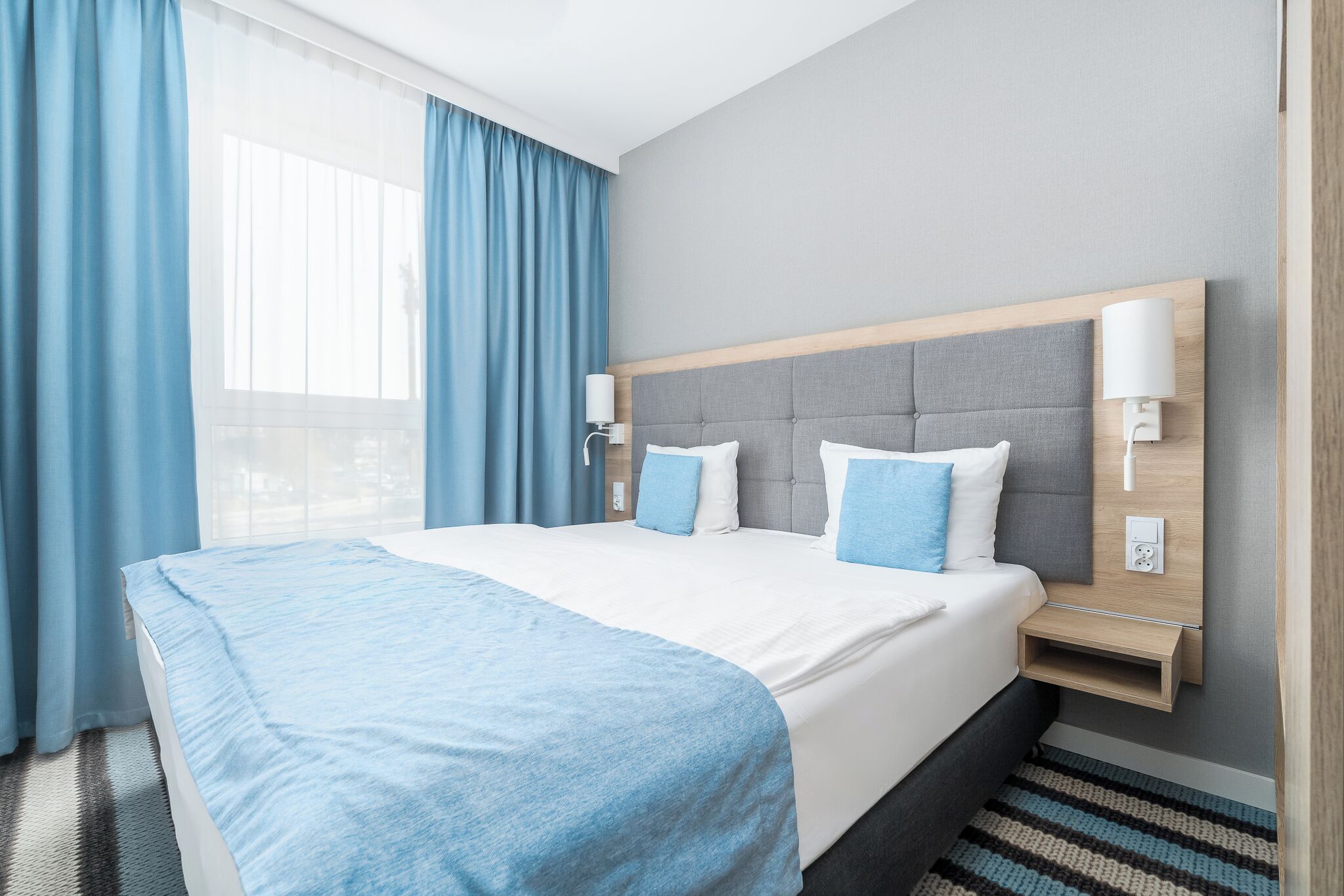 A hotel room with a large bed, two bedside tables, a headboard, and a window with curtains.