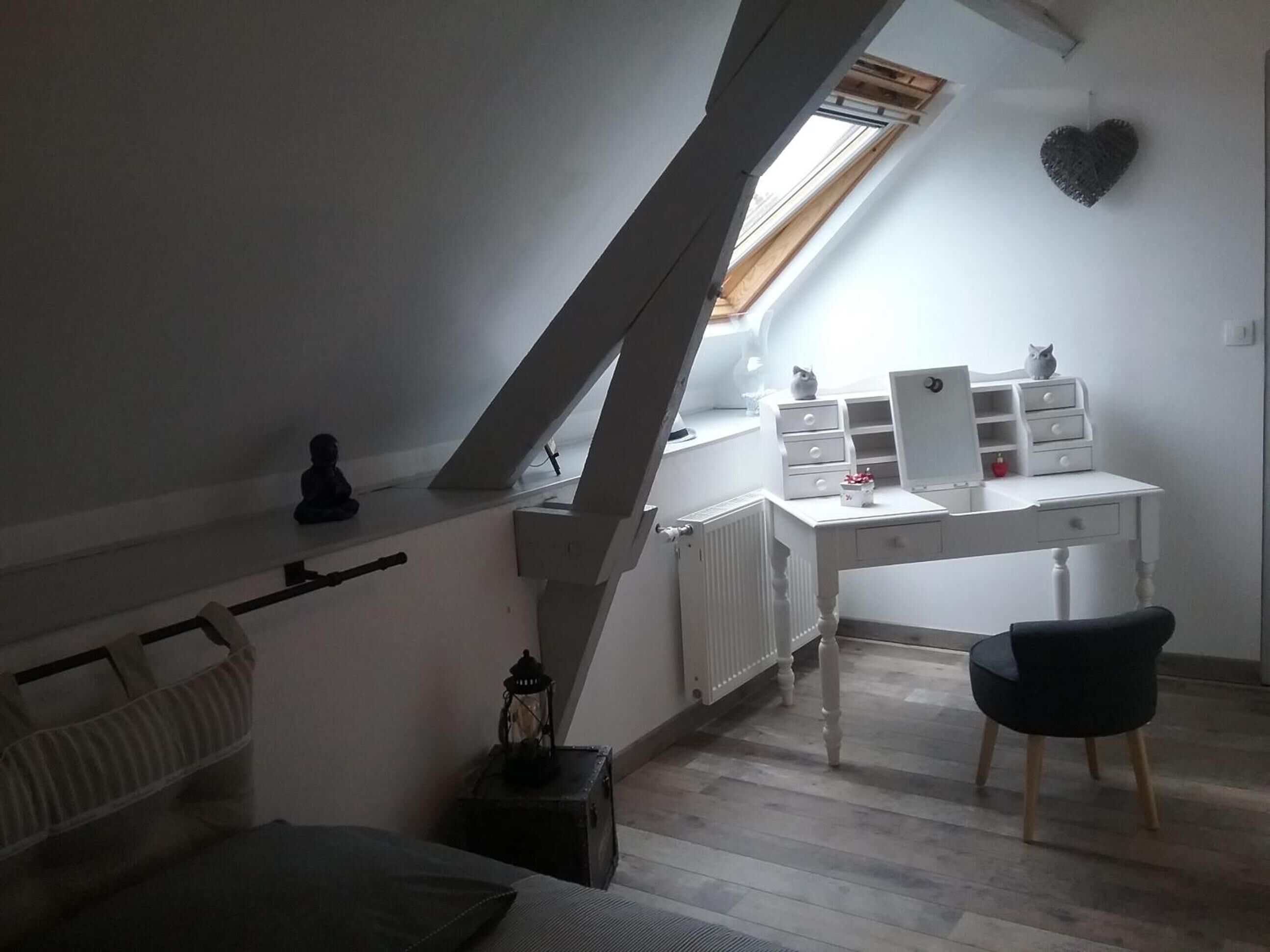 A bedroom with a sloped ceiling, a white desk with drawers, a chair, a radiator, and a window with a view.