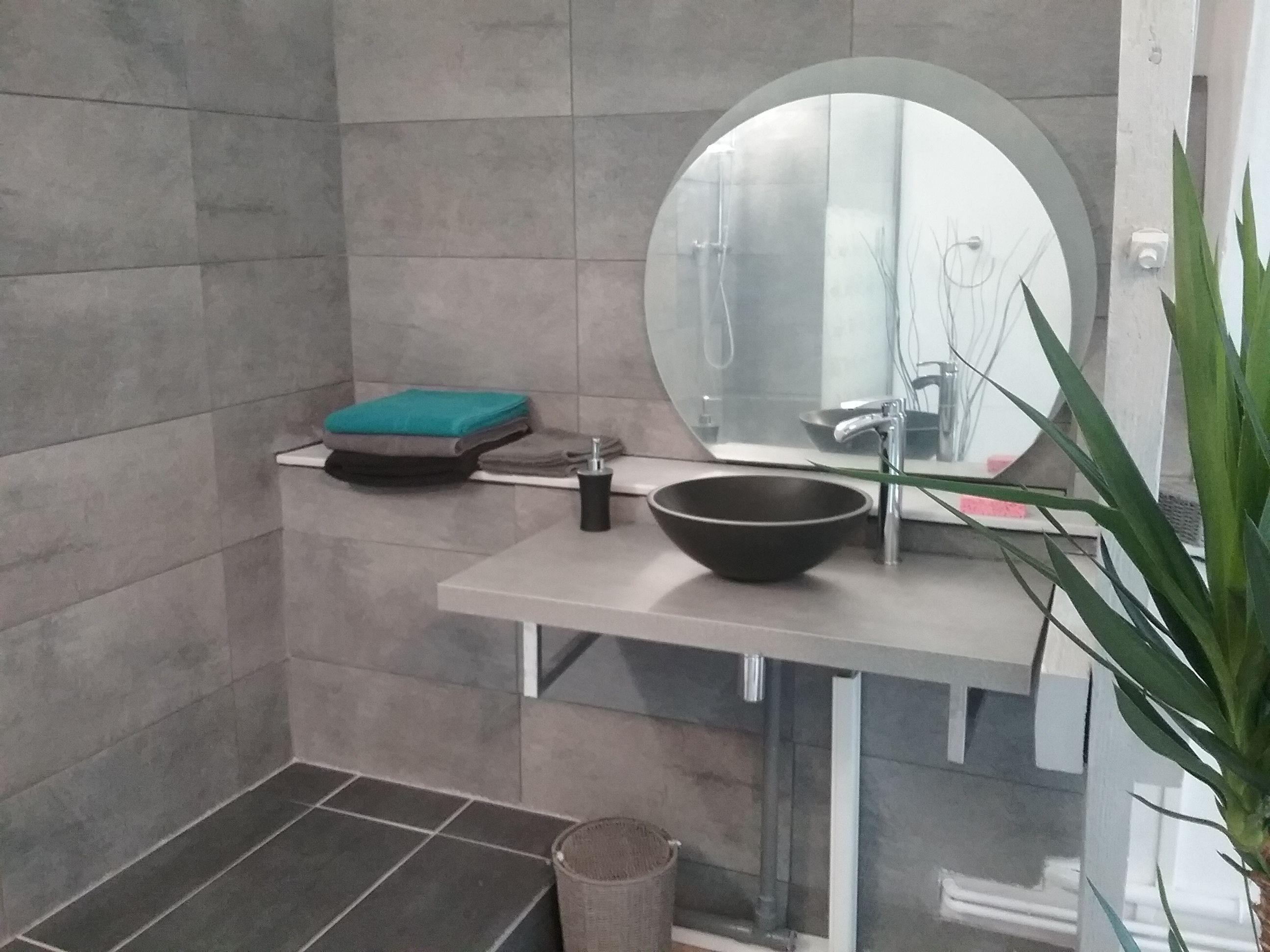 A bathroom with a round mirror, a black bowl sink, a green towel, and a plant.