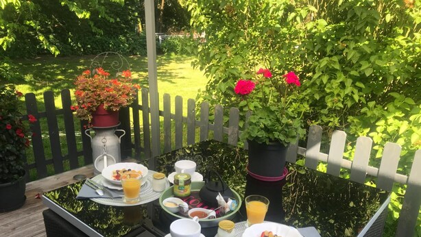 A table set outdoors with breakfast items, surrounded by greenery and a wooden fence. - Chambre d'hôtes Coquelicot (Nurlu)