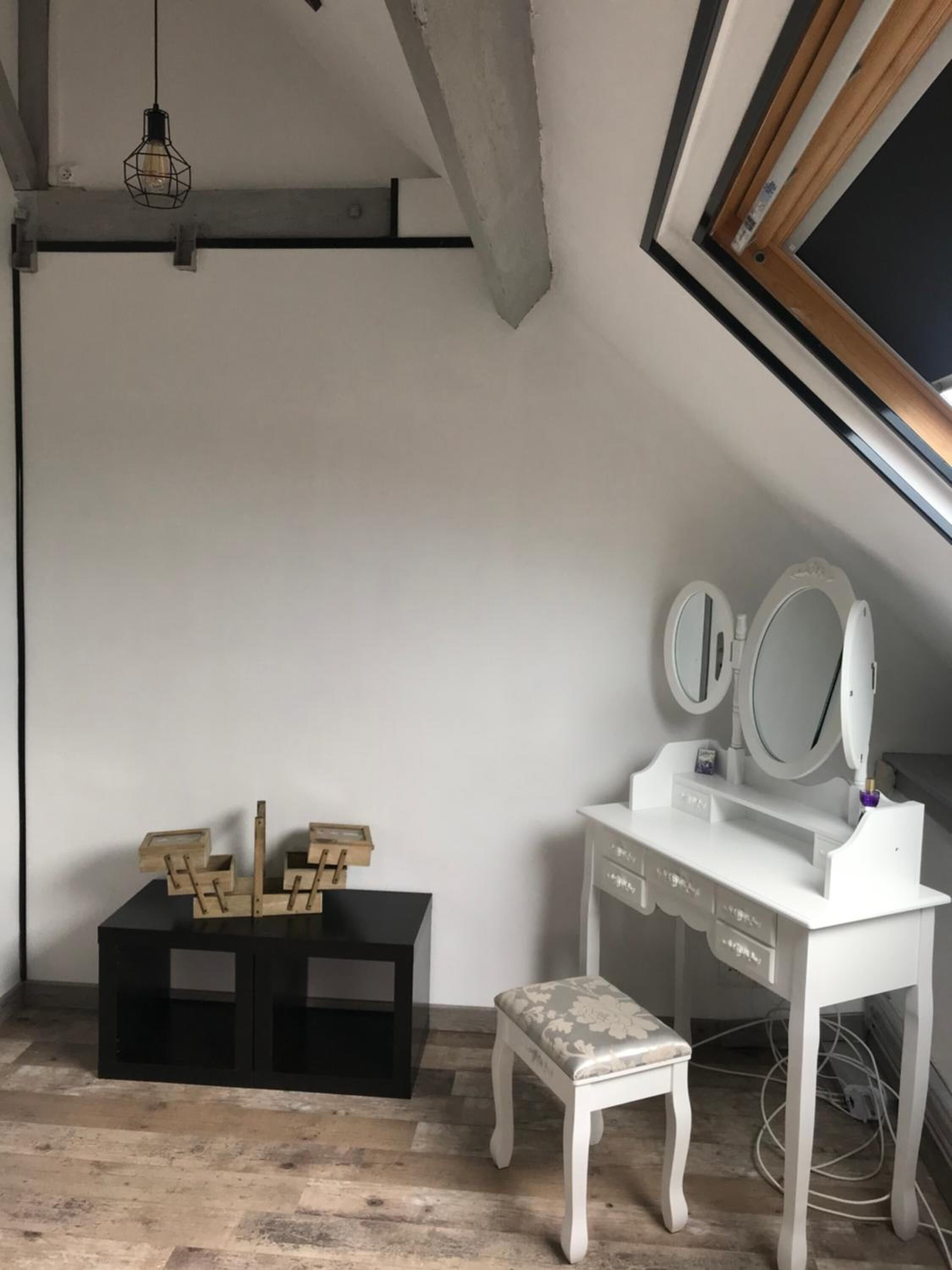 A white vanity with a mirror and a bench, wooden crates, and a hanging light fixture in a room with exposed beams and a sloped ceiling.