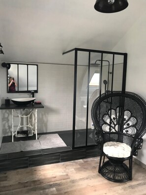 Un baño moderno con cabina de ducha de marco negro, lavabo blanco y silla de mimbre.