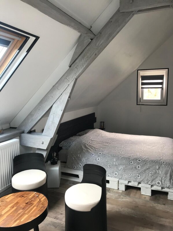 A bedroom with a sloped ceiling, a bed with a floral bedspread, a small table, two chairs, and a window with blinds.