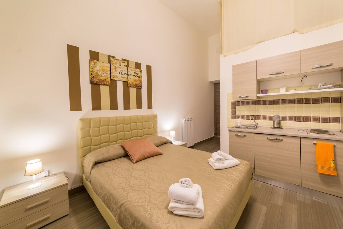 A hotel room with a bed, bedside table, and a kitchenette.