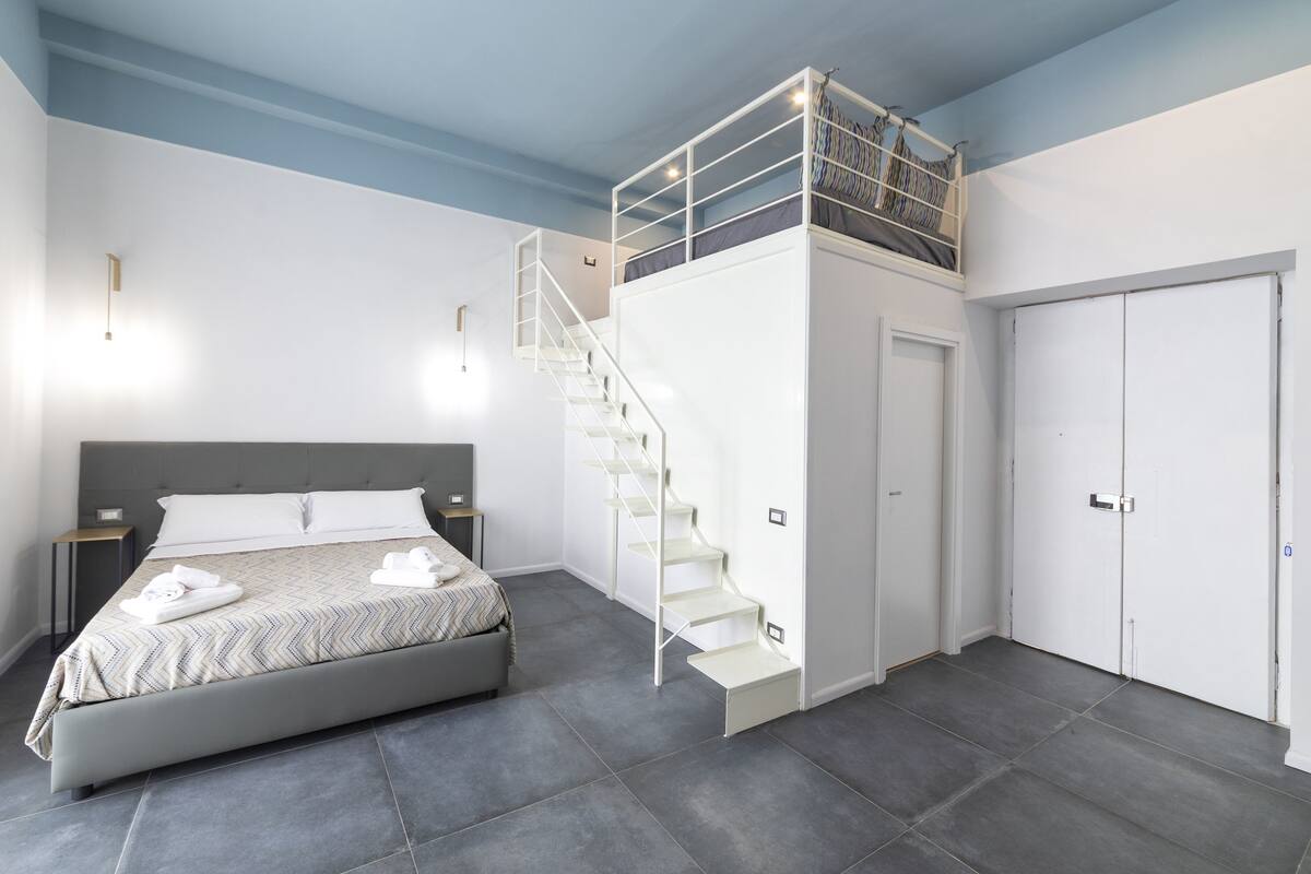 A modern bedroom with a bed, a built-in desk, a staircase, and a wardrobe.