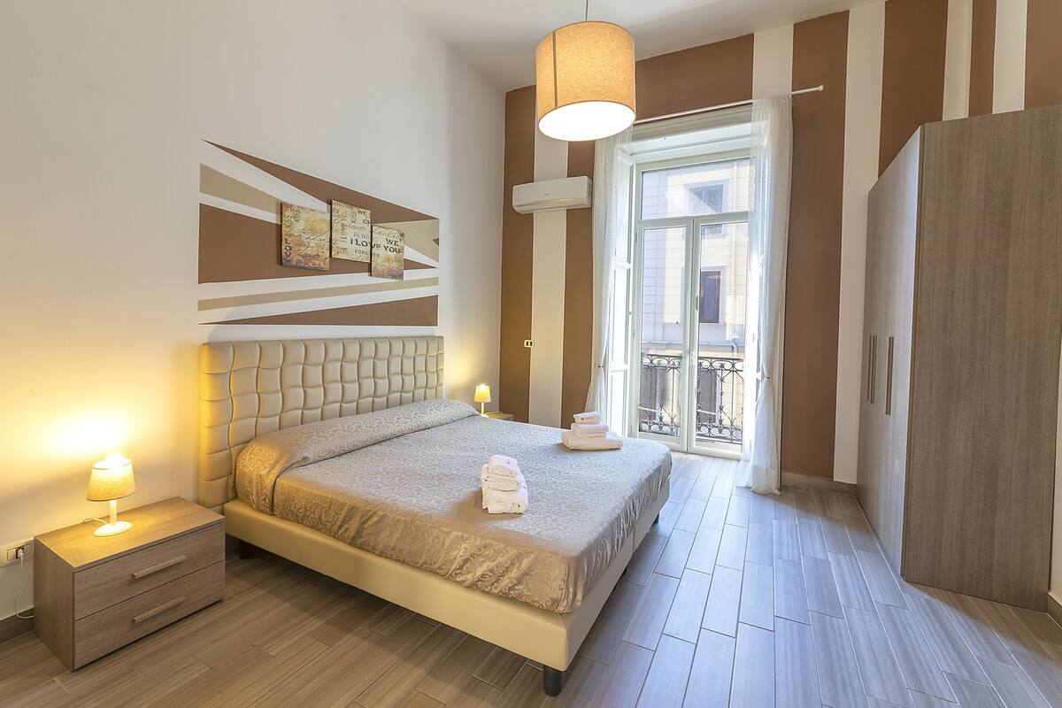A bedroom with a bed, bedside table, lamp, and a view of the outside balcony.