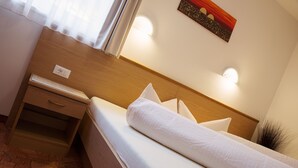 Un lit bien fait, avec du linge de lit blanc et des oreillers, une table de chevet en bois dotée d’un tiroir, et un luminaire fixé au mur.