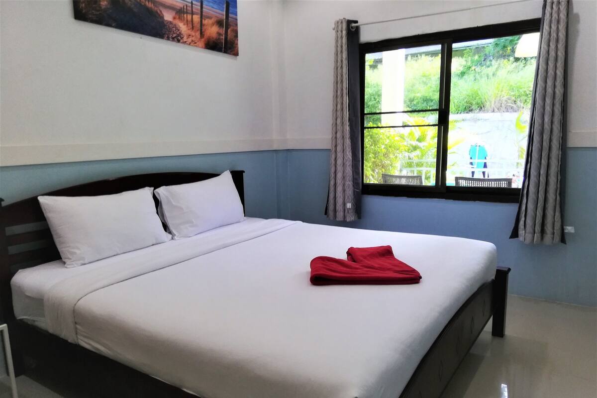 Ein ordentlich bezogenes Bett mit weißen Leinentüchern und einem roten Handtuch, ein Fenster mit Vorhängen und ein Gemälde an der Wand.