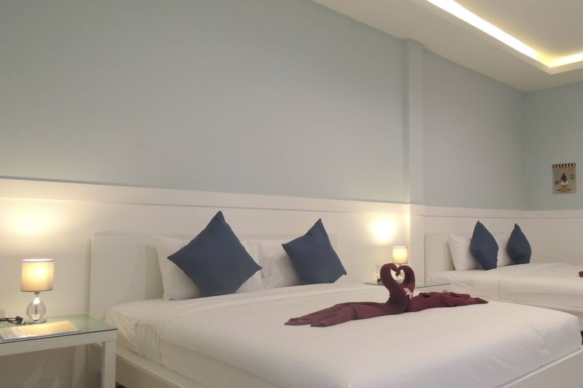 Ein Hotelzimmer mit zwei Betten, jeweils mit weißer Bettwäsche und blauen Kissen. An den Betten befinden sich Nachttischlampen, ein Glastisch und Wandbilder mit Dessertmotiven.