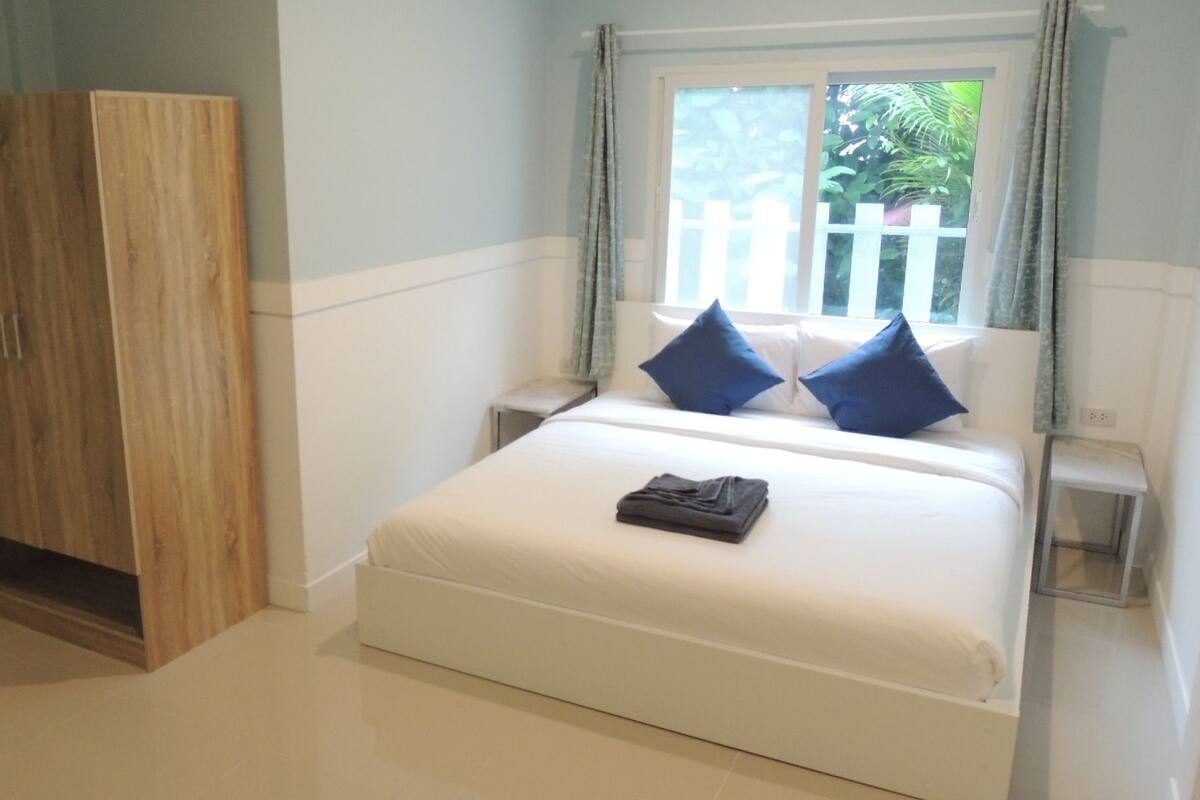 Ein Schlafzimmer mit einem Bett, zwei Kissen, einem Holzschrank und einem Fenster mit Blick auf Grünflächen.