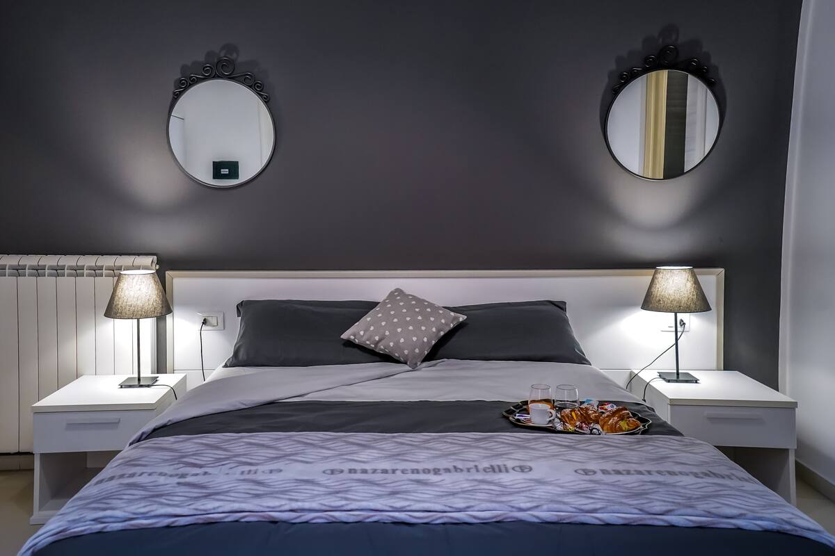 Un dormitorio ordenado con una cama, dos mesitas de noche, un revistero, un espejo y una bandeja con aperitivos.