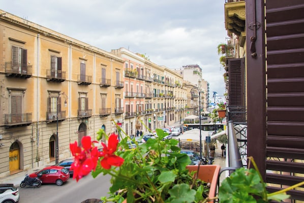 Vista da rua com edifícios, carros e uma varanda com plantas em vasos.