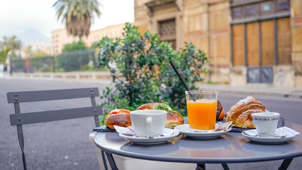 A table with breakfast items including croissants, a cup, and a glass of orange juice, set against a backdrop of a building and greenery. - Amarillo (Palermo)