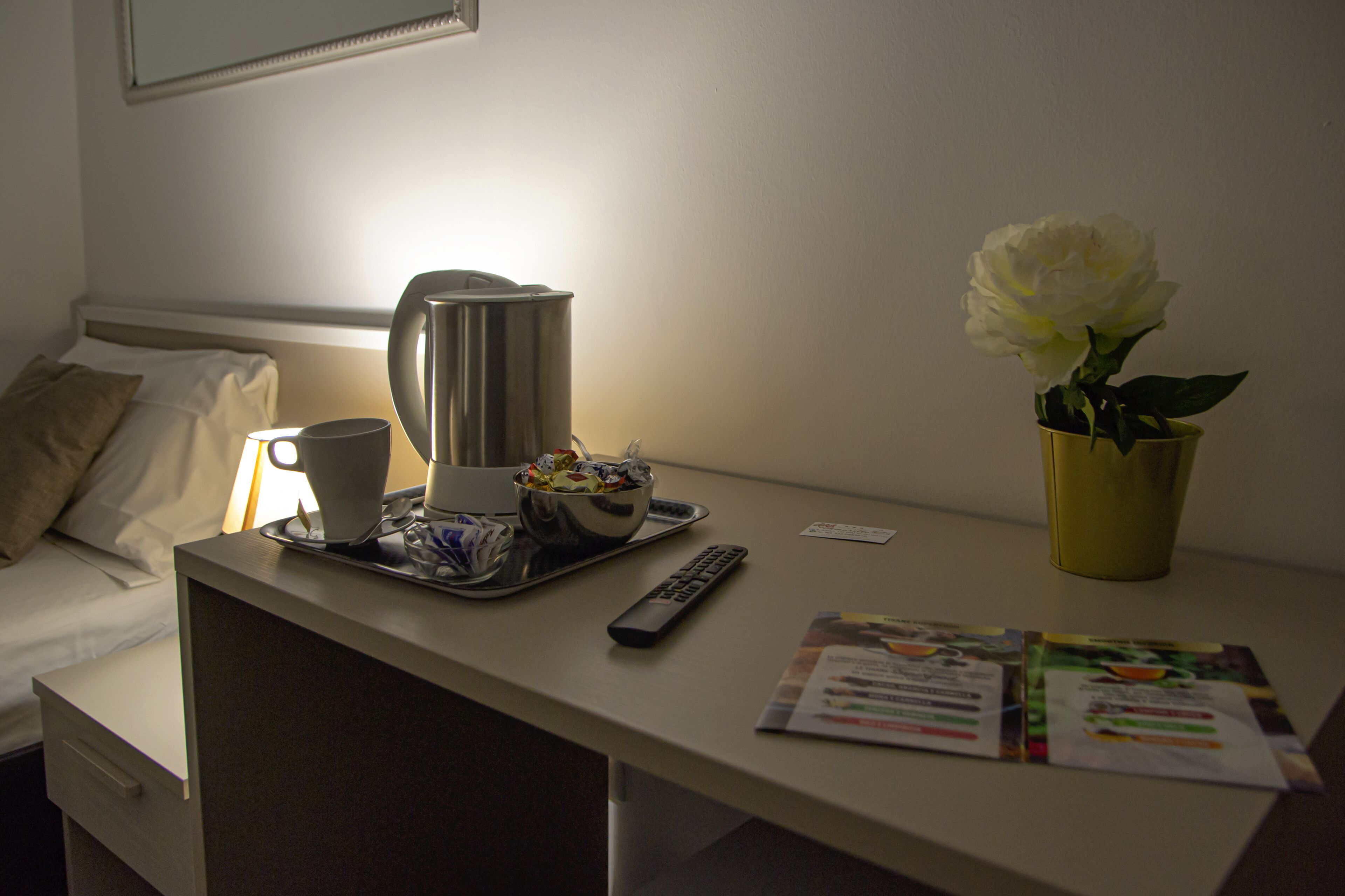 A hotel room with a bed, a bedside table with a coffee maker, a bowl of snacks, a remote control, a magazine, and a vase with a single white flower.