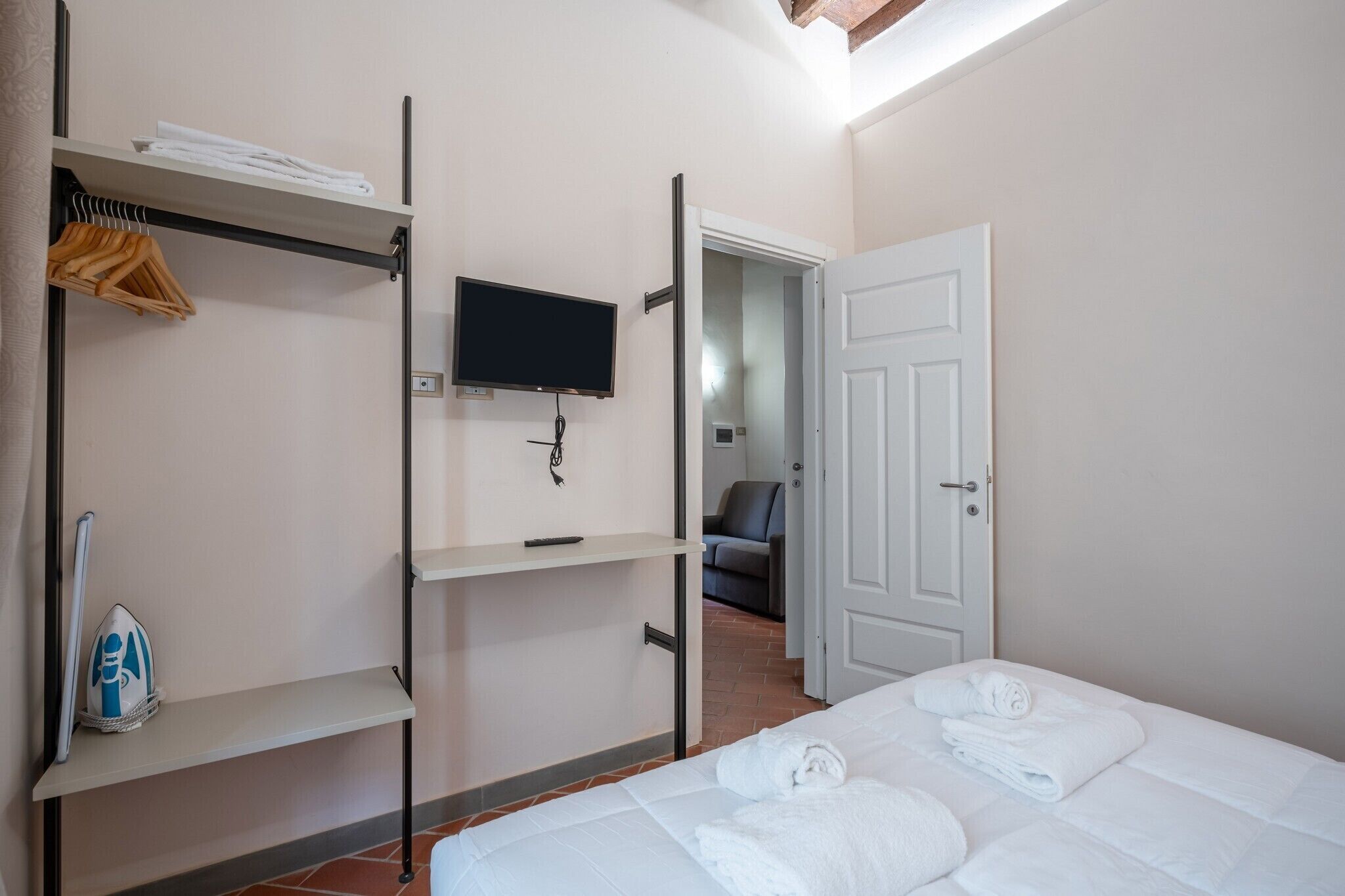A hotel room with a bed, a TV mounted on the wall, a wardrobe with hanging clothes, and a door leading to another room.
