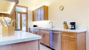 A modern kitchen with wooden cabinets, a white countertop, a built-in dishwasher, and a window with a view of trees.