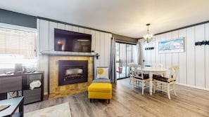 A modern living room with a fireplace, a dining area with a table and chairs, and a large window with blinds. - Mammoth Ski & Racquet Club #4 1 Bedroom Condo by RedAwning (Mammoth Lakes)