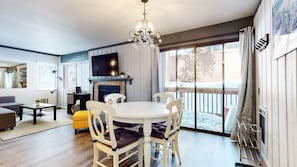 A modern living room with a fireplace, a dining area with white chairs, and a large window offering a view of the outdoors. - Mammoth Ski & Racquet Club #4 1 Bedroom Condo by RedAwning (Mammoth Lakes)