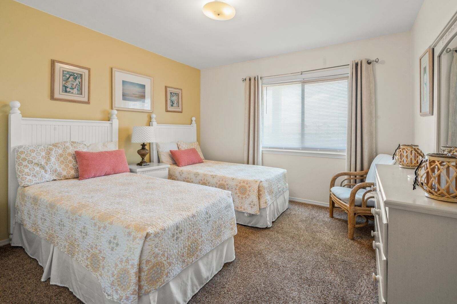 Une chambre avec deux lits, un bureau, une chaise, une fenêtre avec des stores et des tableaux encadrés au mur.