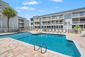 Un espace piscine avec une piscine à l’eau d’un bleu limpide, des chaises longues blanches et un immeuble à plusieurs étages doté de balcons.