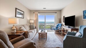 A living room with a sofa, armchairs, a coffee table, a TV, and a view of the sea through a large window. - Sea Cloisters 303e 2 Bedroom Condo by RedAwning (North Myrtle Beach)