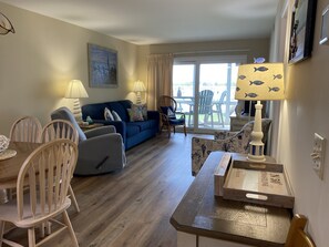 A living room with a blue sofa, wooden chairs, a dining table, and a view through a glass door to the outside. - Sea Cloisters 102b 2 Bedroom Condo by RedAwning (North Myrtle Beach)