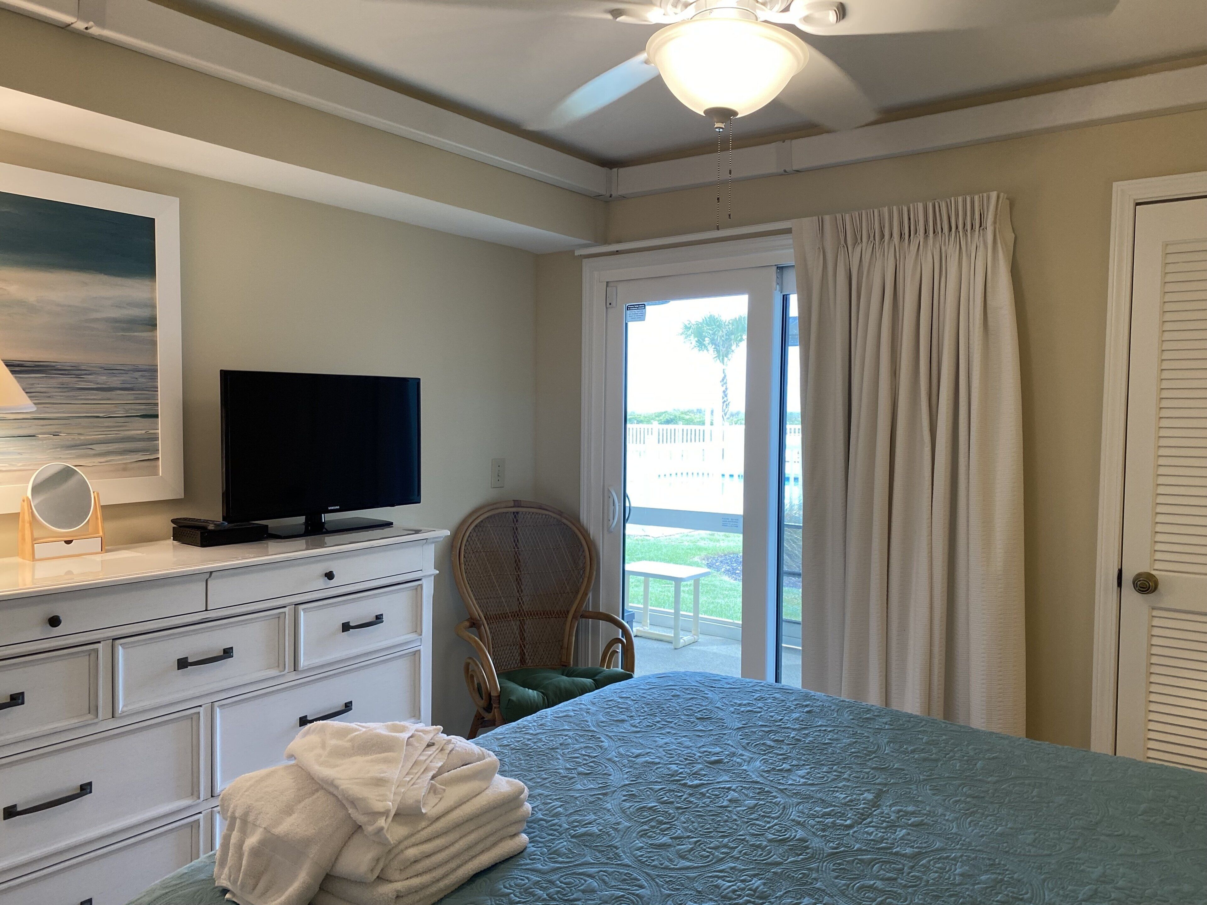A bedroom with a bed, a television on a dresser, a chair, and a view of the outdoors through a door.