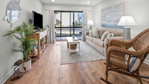 A living room with a TV, sofa, wicker chair, and a large window. - Sea Cloisters 102f 2 Bedroom Condo (North Myrtle Beach)