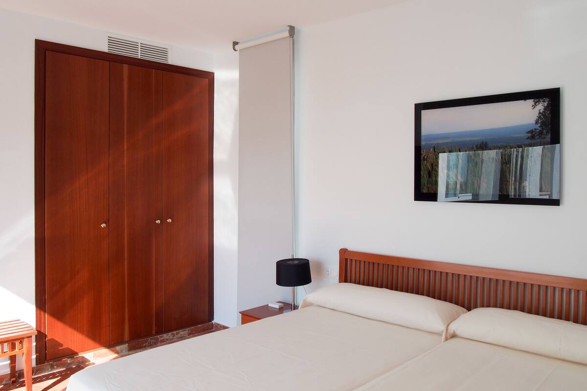 A bedroom with a large wooden wardrobe, a bed with a wooden headboard, a wall-mounted picture, and a small table with a lamp.