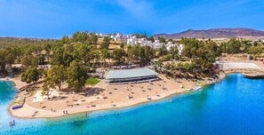 An aerial view of a coastal resort with a beach, a large building, and a mountainous backdrop.