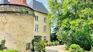 A historic stone building with a round tower, a paved courtyard, and lush greenery.